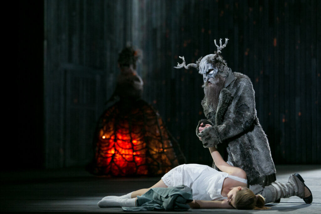 Eine darstellende Person im Rentier-Kostüm hält kniend die Hand einer weiteren liegenden Figur in Weiß