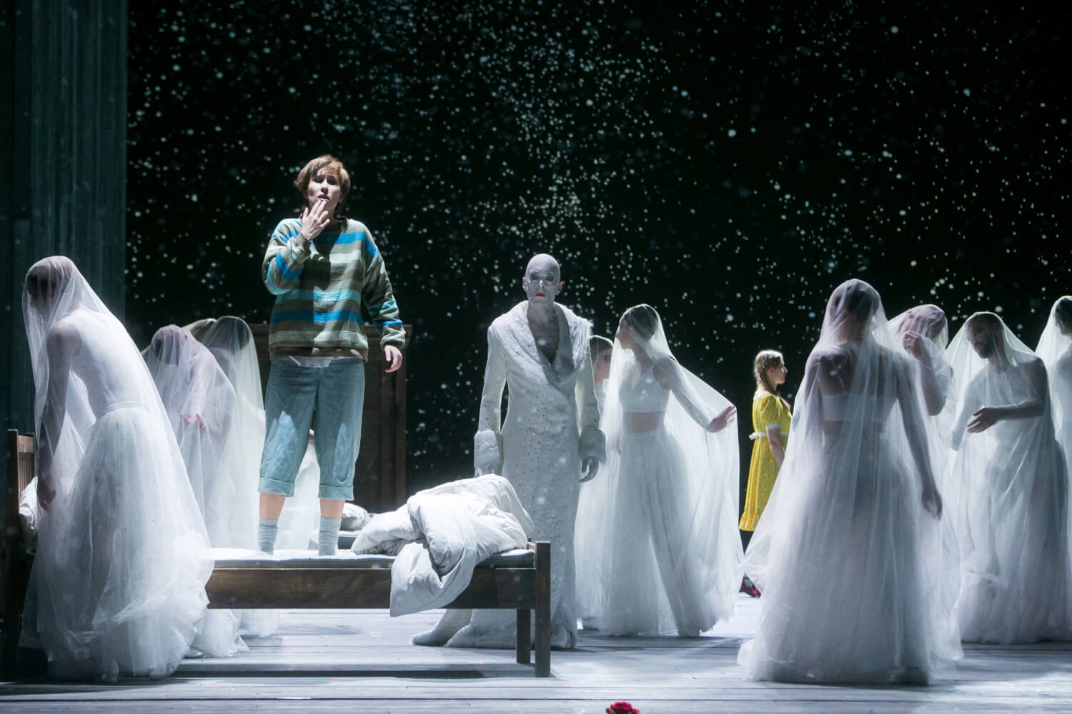 in der Mitte der Bühne eine Bank, auf der eine Person im gestreiften Pulli steht, darum herum weißgewandete Darstellende