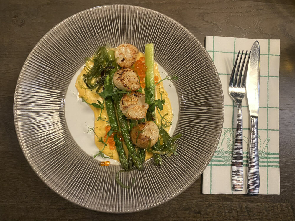 Ein Teller mit drei Stangen grünem Spargel und vier Jakobsmuscheln garniert. Daneben Serviette und Messer und Gabel.