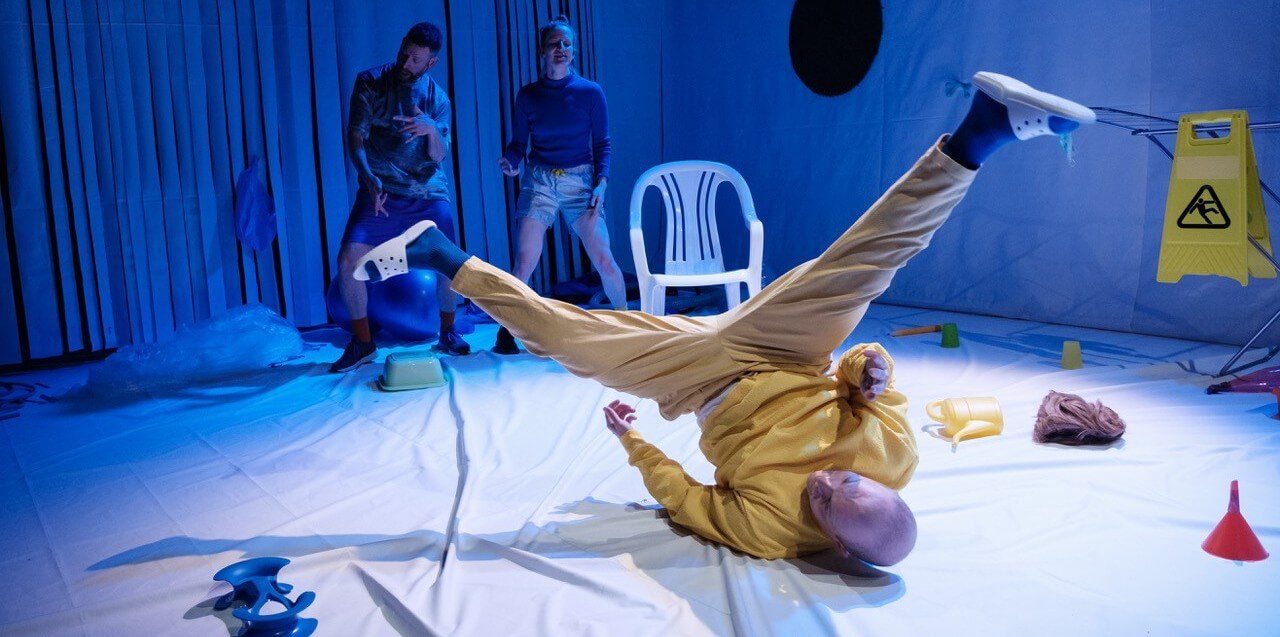 Szenenfoto aus „DINGE DINGEN“. Ein Performer in einem gelben Jogginganzug macht eine Breakdance-Pose. Im Hintergrund steht ein Gartenstuhl und eine Performerin und ein Performer tanzen.