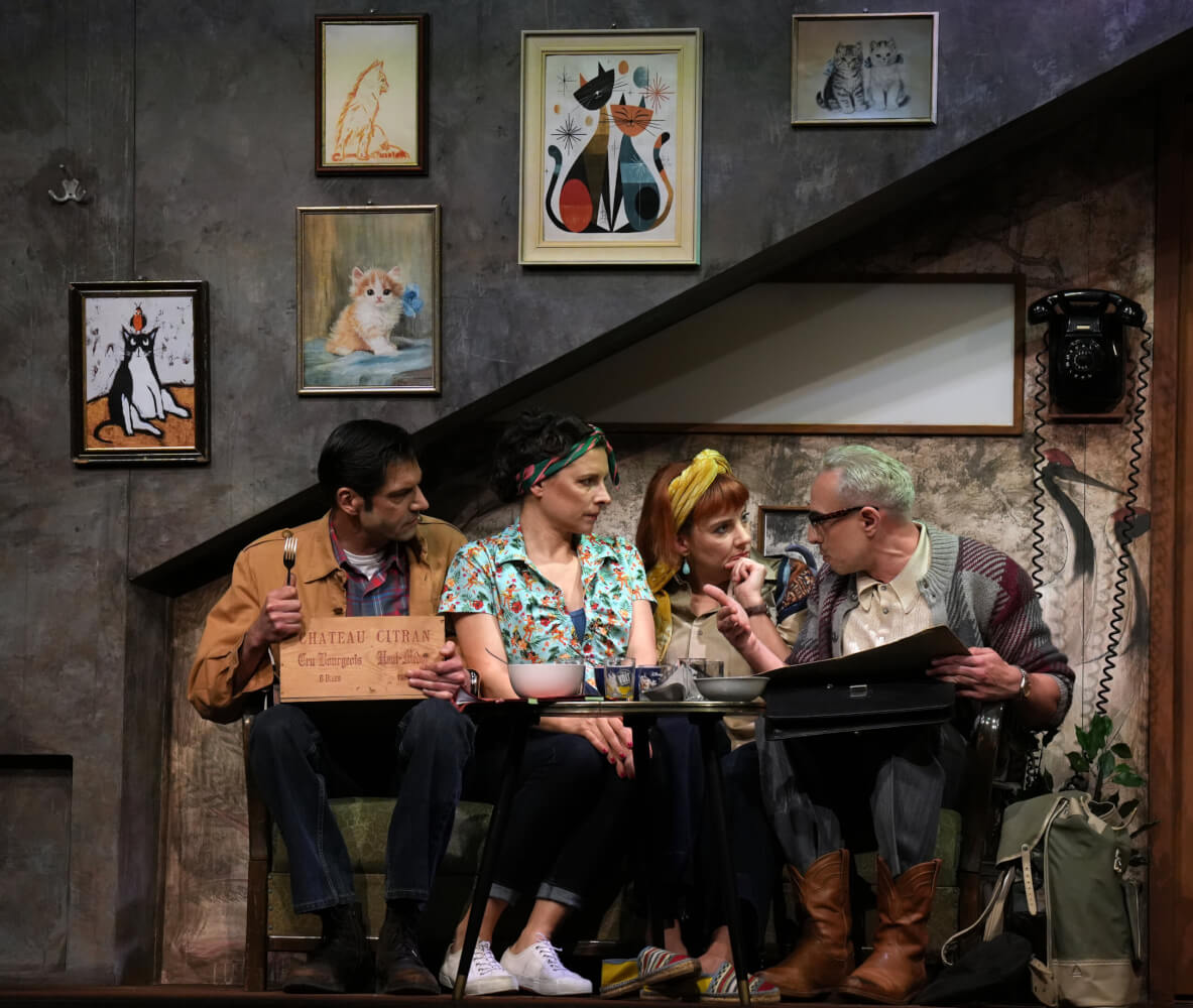 Vier Darstellende sitzen auf der Bühne nebeneinander auf einer Bank und schauen sich an. Sie scheinen Pläne zu schmieden. Im Hintergrund hängen an einer Wand unterschiedliche Bilderrahmen.