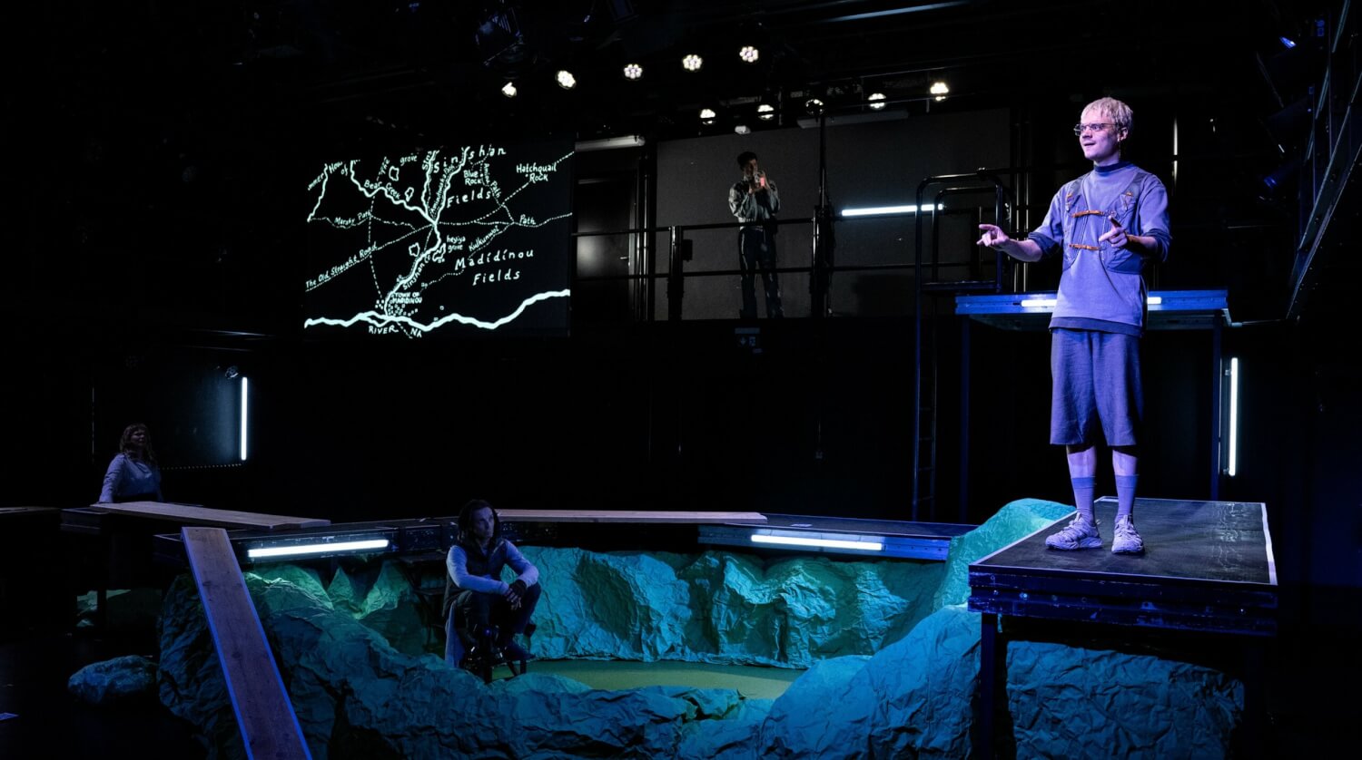 Aufführungsfoto von „Immer nach Hause“ nach Ursula K. Le Guin, von F. Wiesel. Ein grün-blau beleuchteter Krater in der Mitte der Bühne, in dem ein Mann sitzt. Auf dem Podest rechts oben steht ein Mann, blickt und zeigt ins Publikum. Im Hintergrund links eine Leinwand mit hellen Strichen.