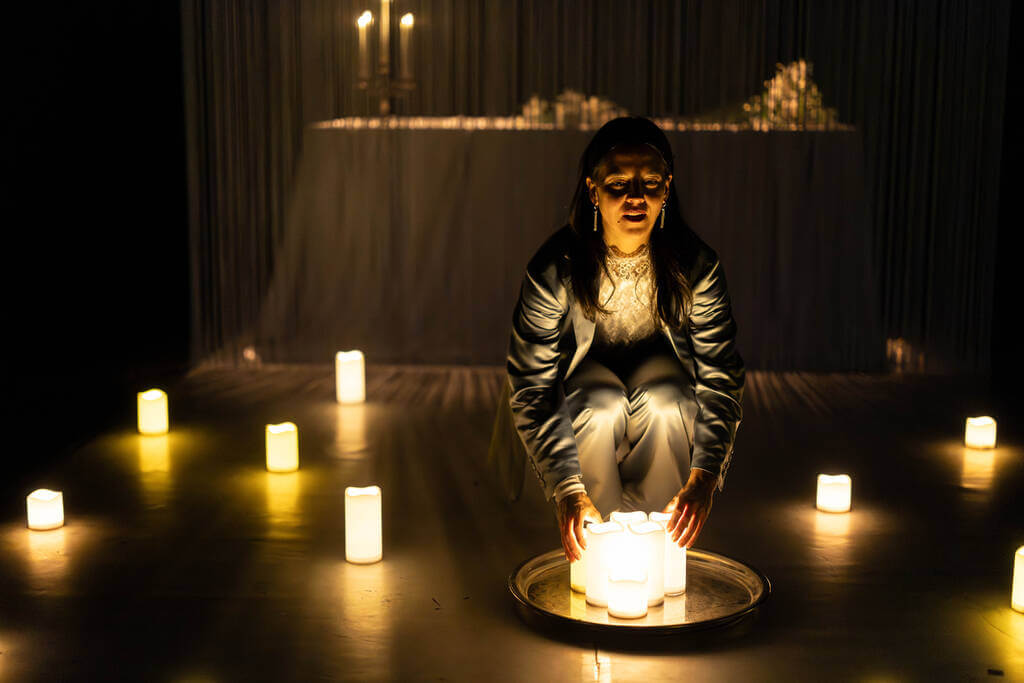 Aufführungsfoto von „Söhne“ von Marine Bachelot Nguyen am Schlosstheater Moers mit Catherine Elsen. Eine Frau hockt in einem dunklen Raum auf dem Boden und hält eine Gruppe von Kerzen in den Händen. Um sie herum stehen verstreut ebenfalls erleuchtete Kerzen auf dem Boden.
