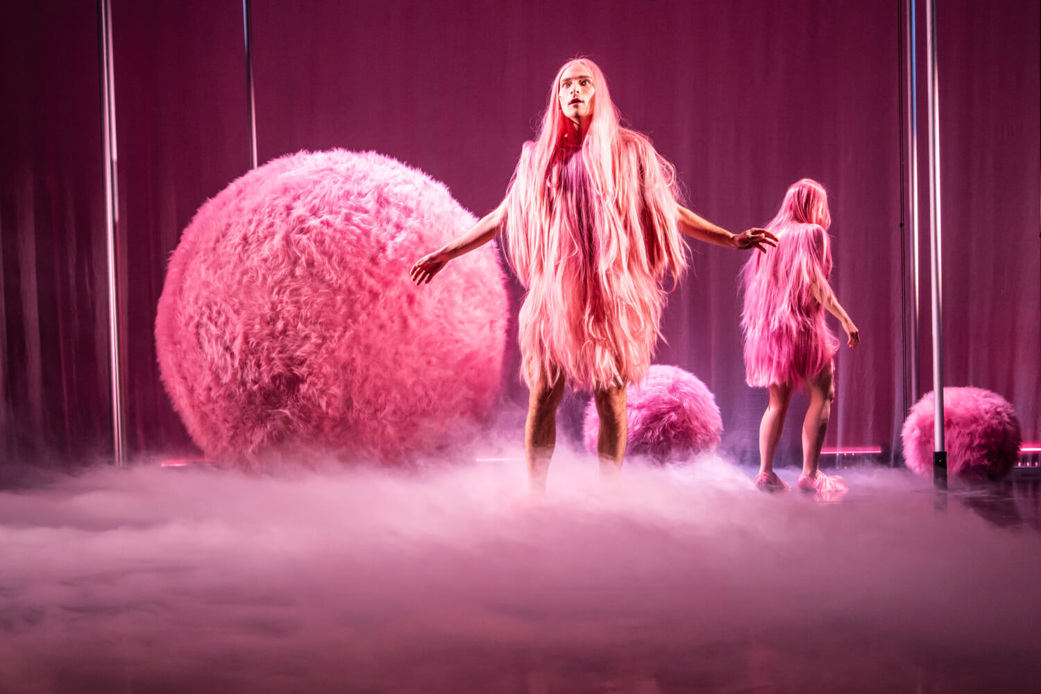 „Die Hölle auf Erden“ von Maria Lazar am Theater Magdeburg. Ein Mann steht im Bühnenbodennebel vor drei unterschiedlich großen rosa Plüschkugeln. Er selbst trägt eine rosafarbene Perücke, die in das rosa Langhaarkostüm nahtlos übergeht.