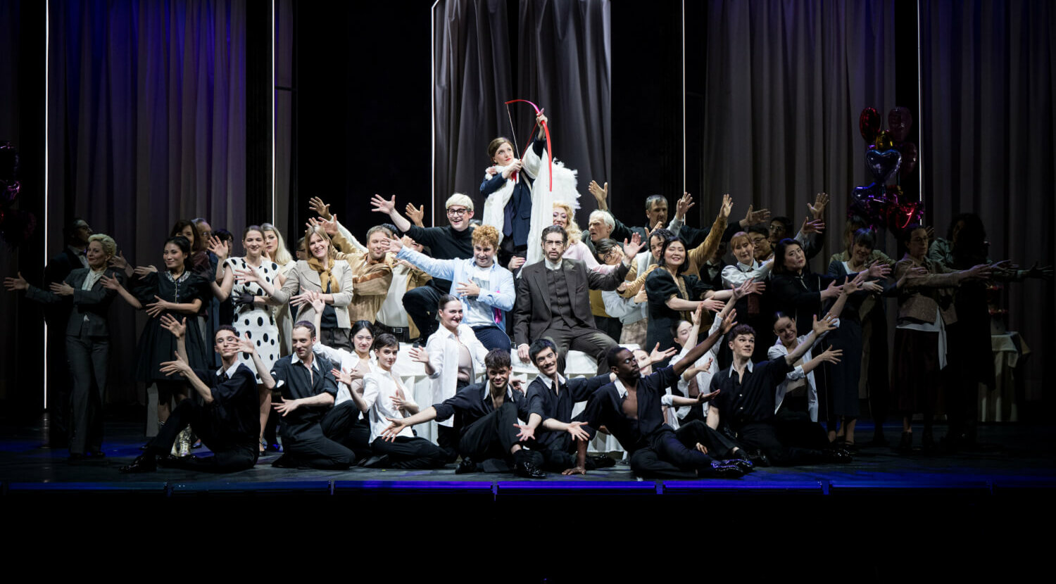 Ensemblebild, Amor mit rotem Pfeil zu erkennen, darum herum mit erhobenen Armen Opernsänger:innen, Chor und Tänzer:innen