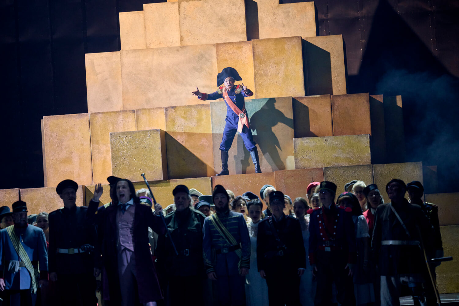 Aufführungsfoto von „Napoleon“ von Armin Petras am Saarländischen Staatstheater. Im Vordergrund eine Menschenmasse im Dunkeln. Im Hintergrund steht Napoleon auf beigefarbenen Quadern.