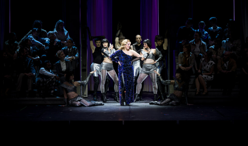 beleuchtet in der Mitte eine Frau im blauen Glitzerkleid, darum herum ein posierendes Ensemble