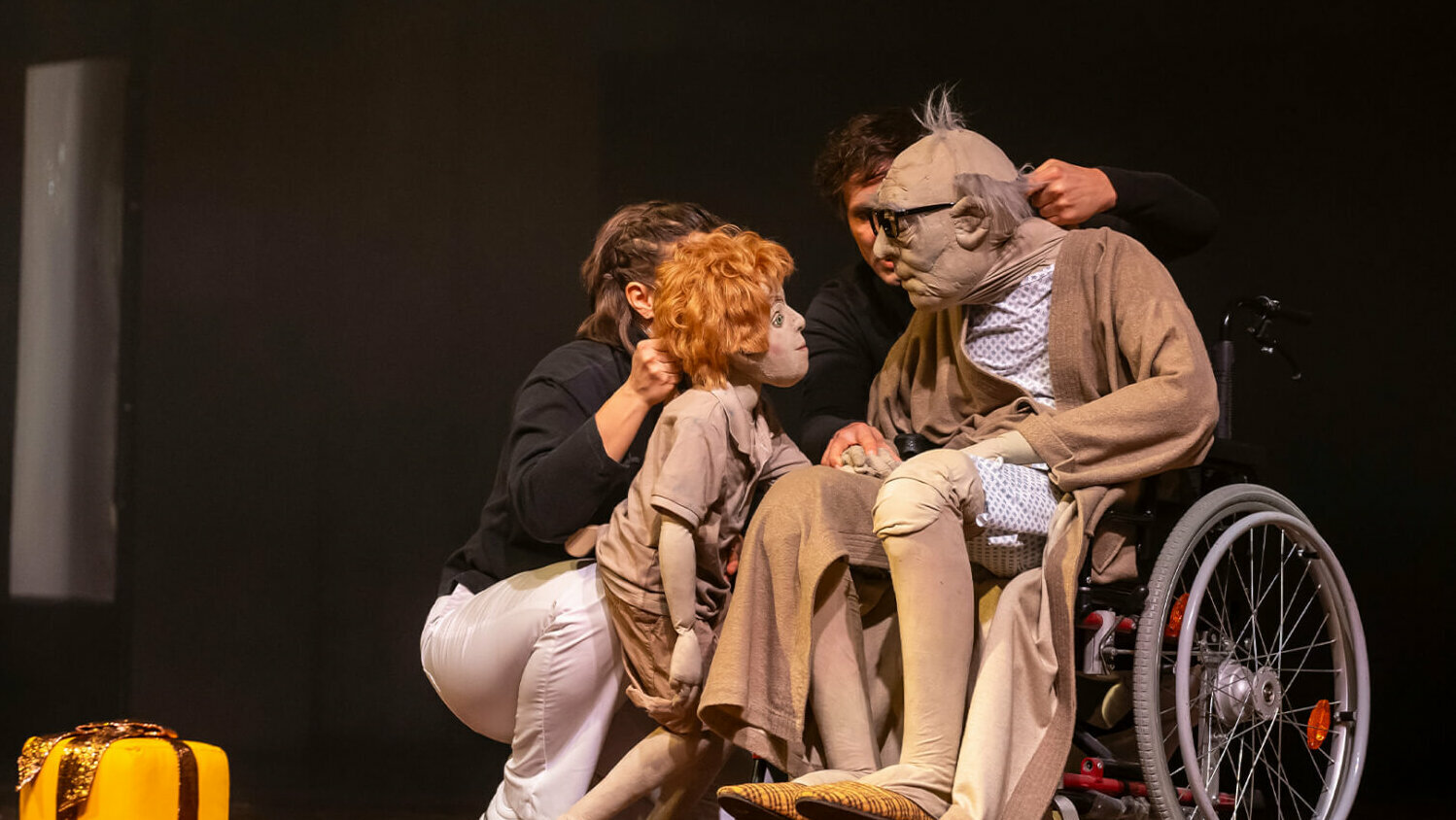 Eine lebensgroße Puppe einer alten Person im Rollstuhl, daneben die eines Kinds mit rotblonden Haaren, dahinter zwei Spieler:innen