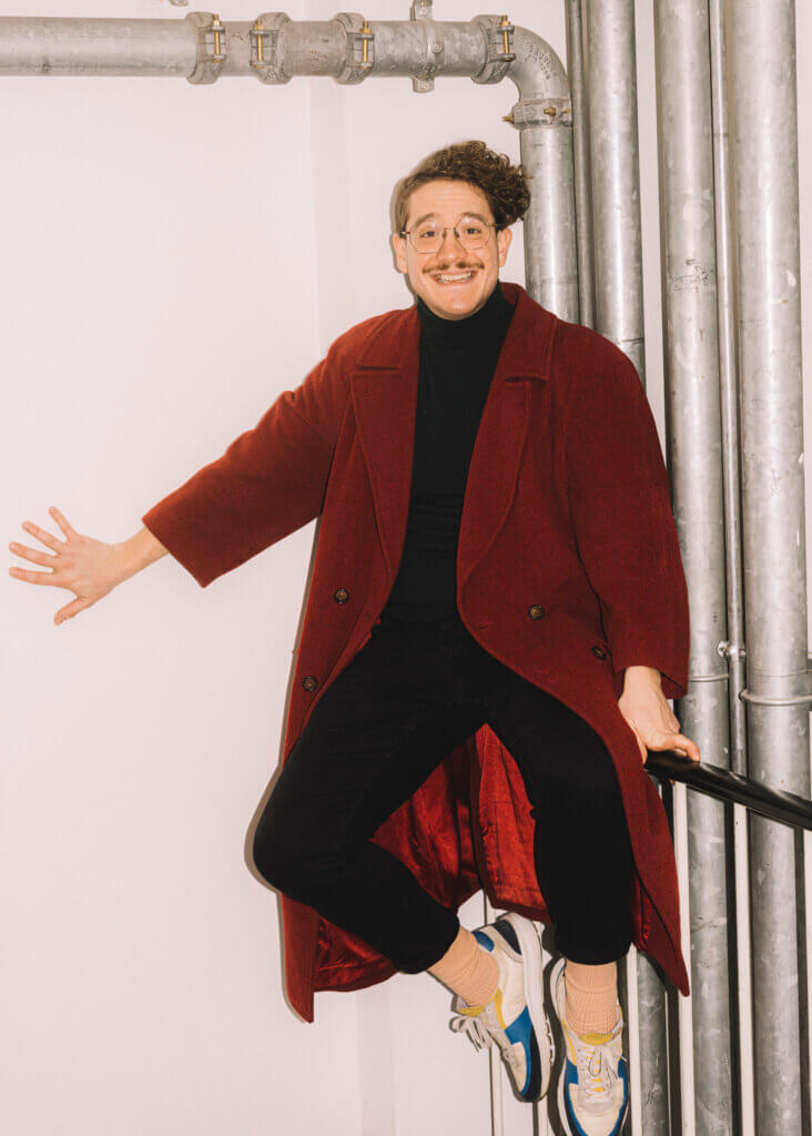 Maurice Lenhard in einer Raumecke, an der vier Rohre entlang laufen, stützt sich mit der linken Hand an einer Stange und mit der rechten Hand an der Wand ab und steht auf Zehenspitzen.