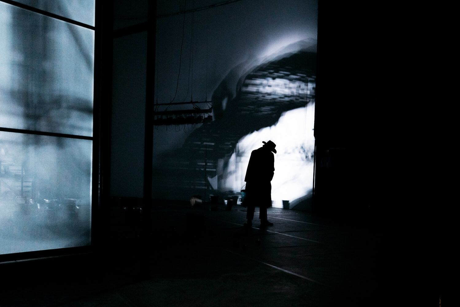 Die Bühne ist so belichtet, dass Nebelschwaden die Wände hochzusteigen scheinen. Im Hintergrund steht eine schattenhafte Person mit Mantel und Hut.