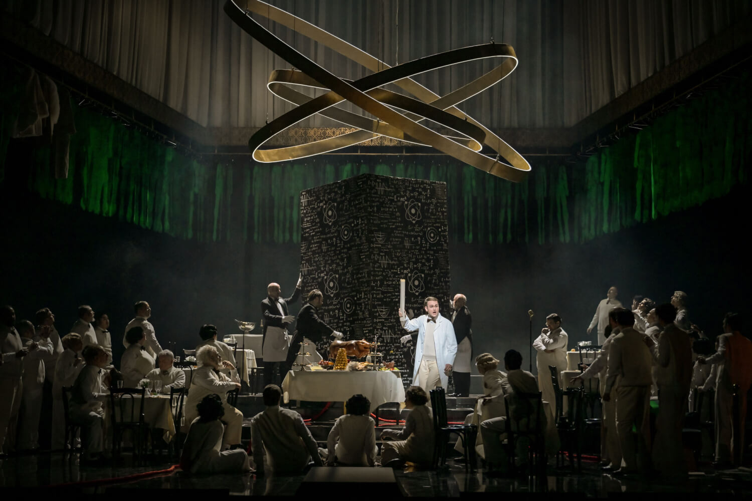 ein astrologisches Gebilde, darunter auf der Bühne Darsteller:innen verkleidet als Wissenschaftler:innen