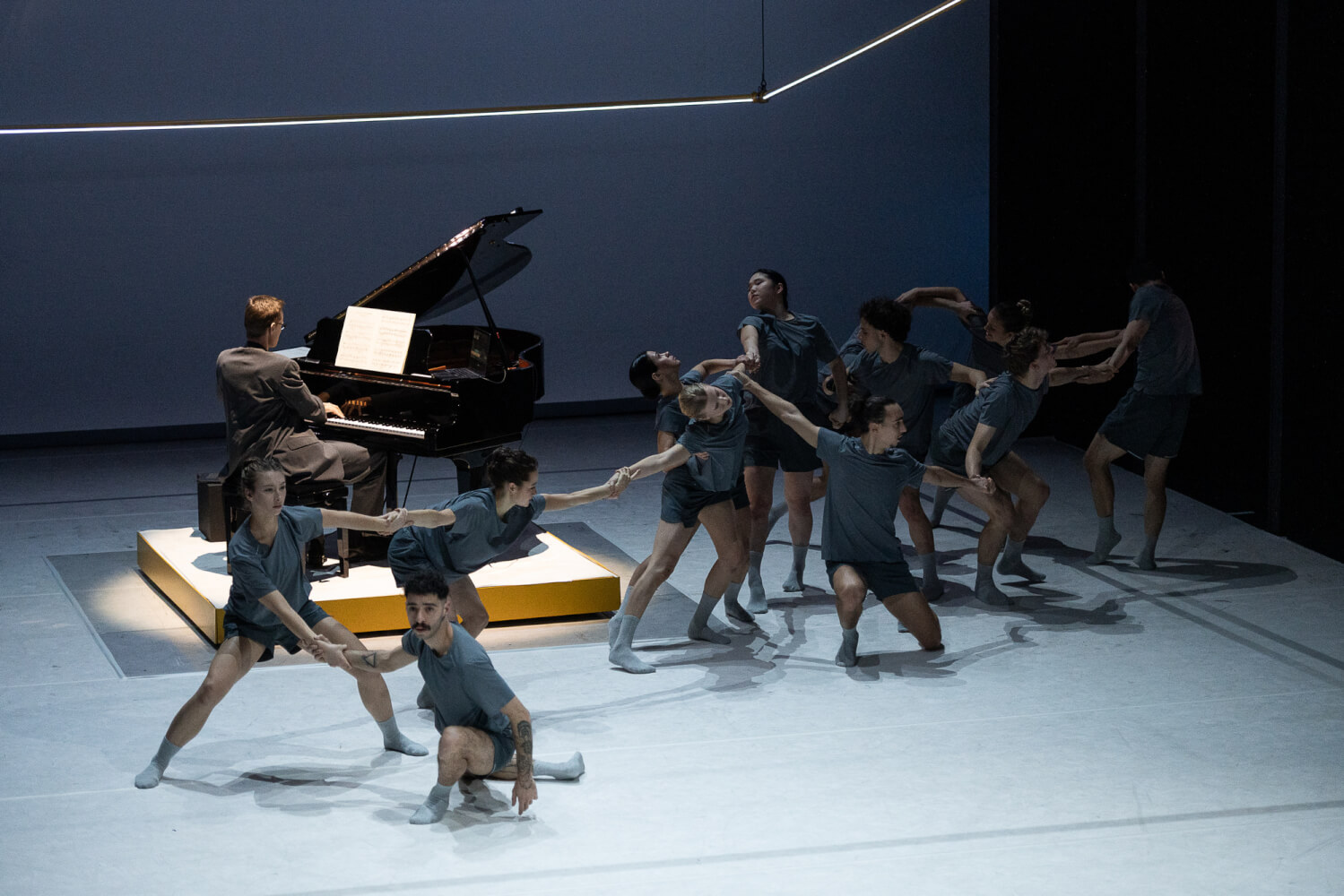 Aufführungsfoto von „UTOPIA? DIY! Eine Utopie“ von Massimo Gerardi am Gerhart-Hauptmann-Theater Görlitz-Zittau. Elf Tänzerinnen und Tänzer mit dunkel grauen T-shirts halten sich wie an einer Kette an den Händen. Dahinter sitzt ein Pianist am Flügel.