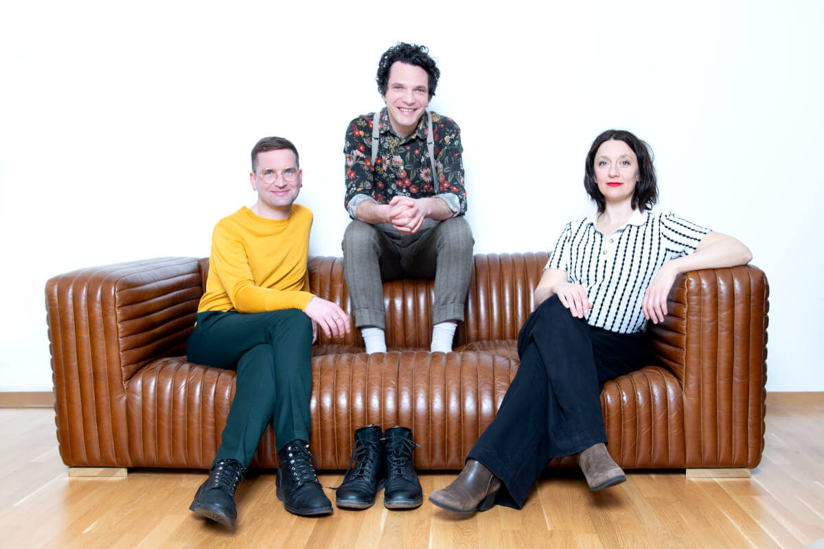 Clemens Leander, Bastian Lomsché und Clara Weyde sitzen auf einer braunen Ledercouch. Bastian Lomsché in der Mitte sitzt auf der Lehne, seine Schuhe stehen auf dem Fußboden.