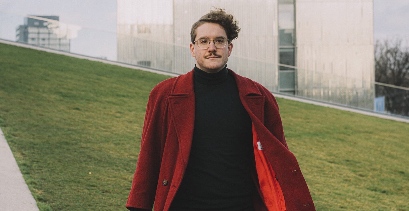 Maurice Lenhard steht vor einem hellen Hochhaus, vor einem grünen Rasen und trägt einen langen roten Mantel.