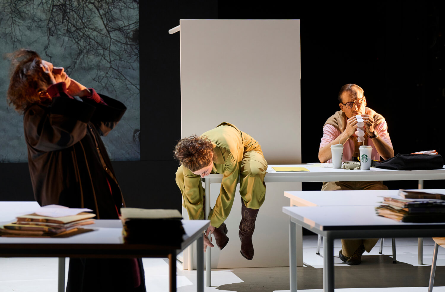 Auf der Bühne stehen Tische wie im Klassenzimmer aufgereiht. Eine Person sitzt mit Kaffee in der letzten Reihe. Eine weitere sitzt auf die Knie gebeugt auf einem Tisch weiter vorne und starrt auf den Boden. Vorne links steht eine Person im langen dunklen Mantel und wirft benommen die Haare zurück.