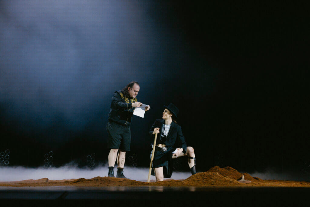 Zwei Darsteller:innen auf einer mit Erde bedeckten Bühne. Links stehend liest von einem Papier, rechts auf einem Bein kniend schaut nach links zur anderen Person