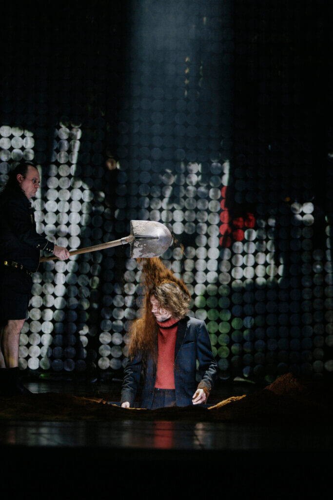 Ein Darsteller in einem ausgehobenen Grab auf der Bühne, ein anderer lässt gerade Erde von einer Schaufel auf dessen Kopf fallen