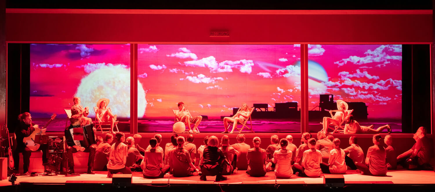 „AscheMOND oder The Fairy Queen“ von Helmut Oehring am Staatstheater Cottbus. Die Bühne ist in orange-pinkes Licht getaucht. Im Hintergrund zwei Planeten und Wolken. Fünf Personen sitzen in Sonnenstühlen. Im Vordergrund eine Menschenmenge, die die fünf Personen ansehen.