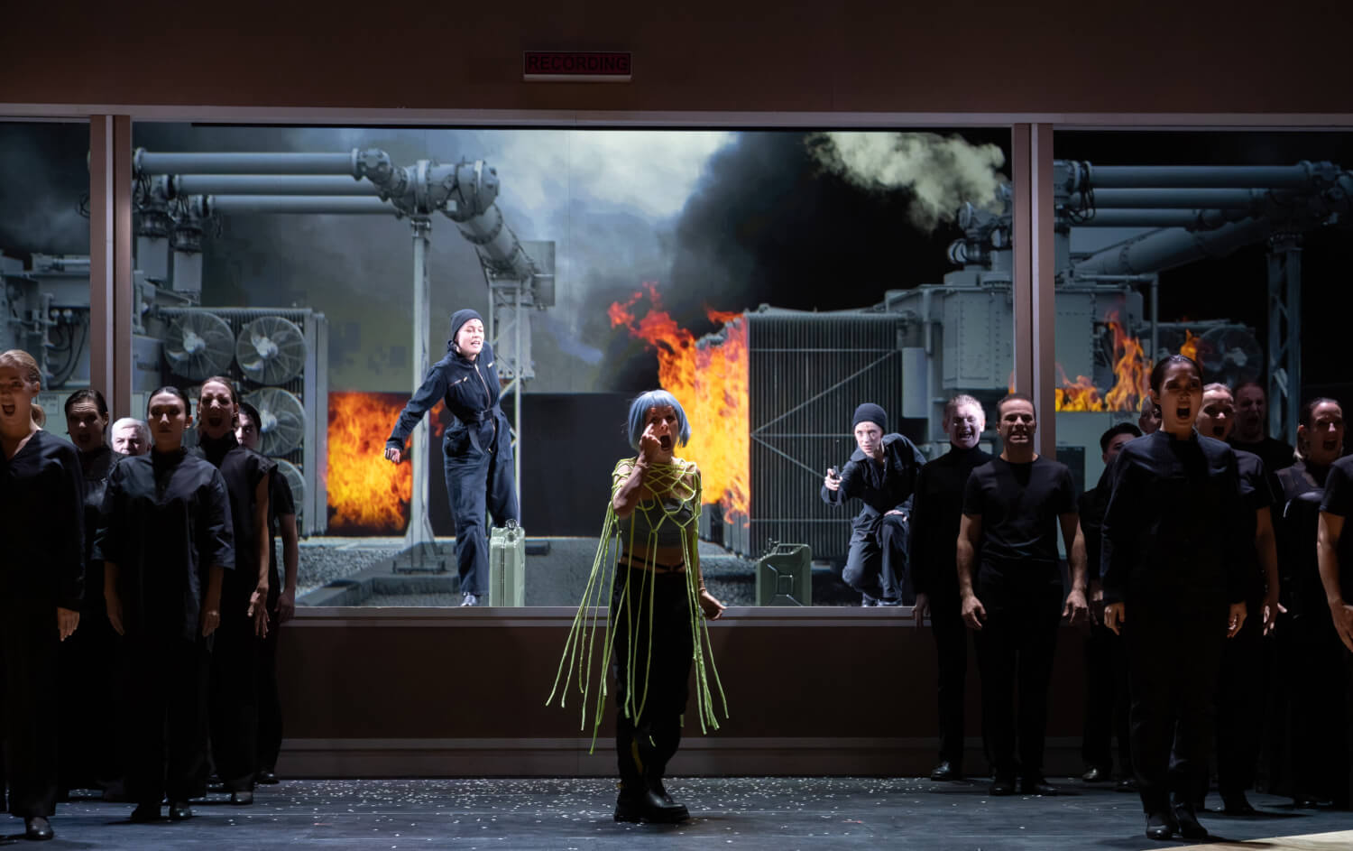 „AscheMOND oder The Fairy Queen“ von Helmut Oehring am Staatstheater Cottbus. Im Hintergrund Flammen und Industrierohre. In der Mitte eine Fraz mit Perücke, die ihren Mund aufreißt. Links und rechts ein Chor, der schwarz angezogen ist.