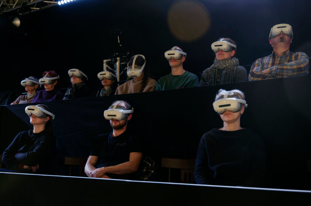 Das Publikum sitzt nebeneinander im dunklen Zuschauerraum. alle haben ihre gesichter nach vorn gewandt und tragen AR-Brillen.