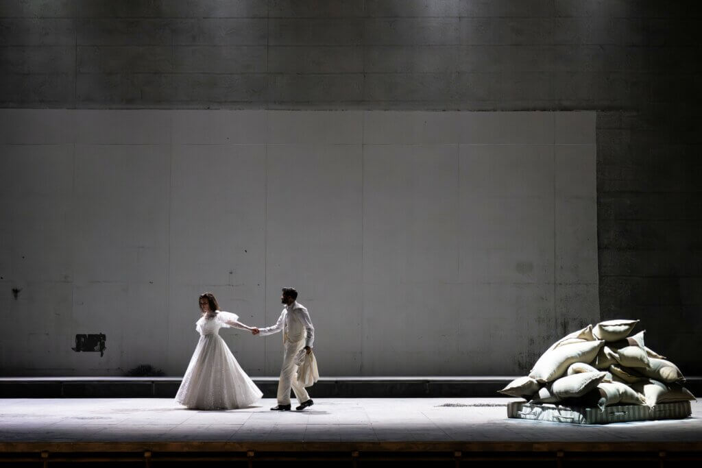Zwei Darstellende stehen links auf der Bühne. Rechts liegt ein Haufen aus Decken. Ansonsten ist die Bühne leergefegt.