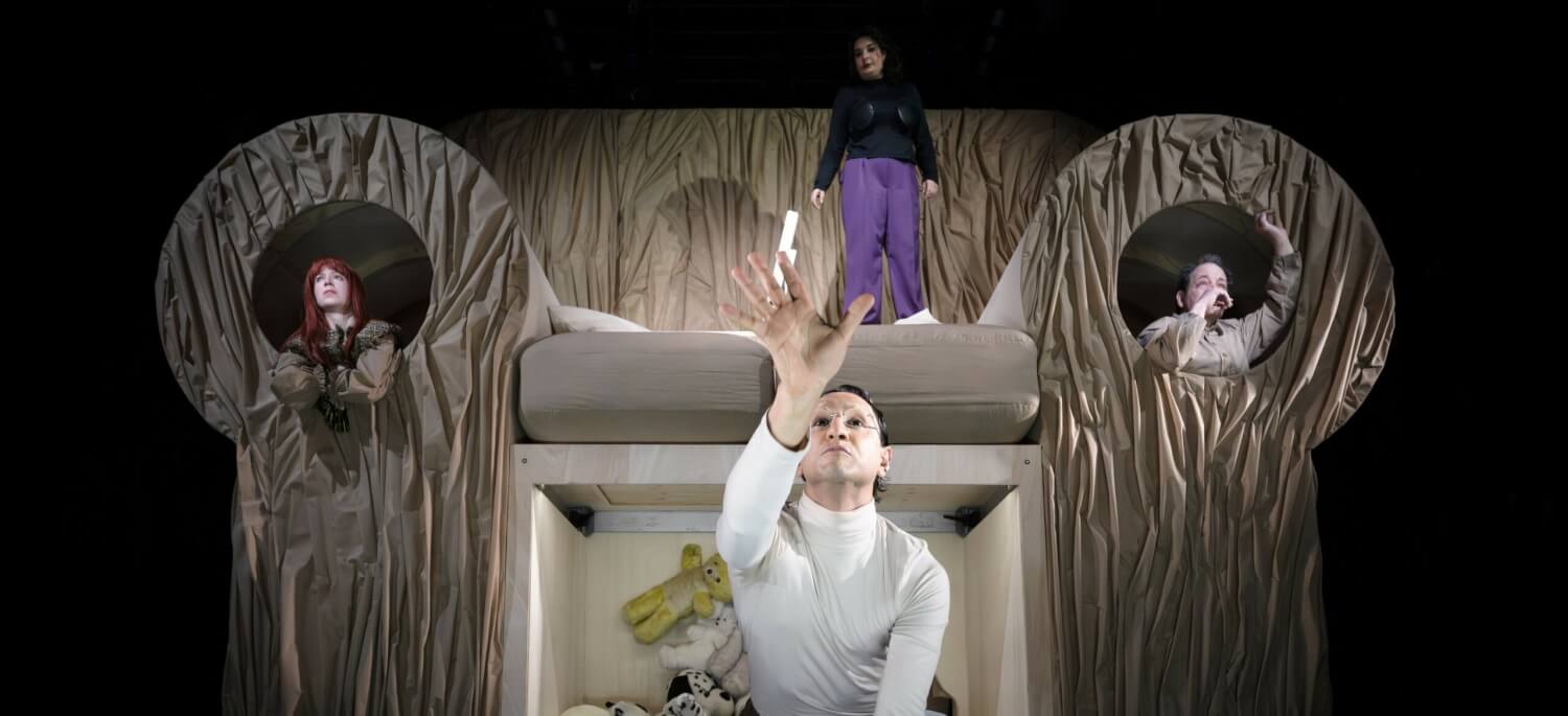 Aufführungsfoto von „Es sagt, es liebt uns“ von Emre Akal am Nationaltheater Mannheim. Im Hintergrund ein mit Packpapier verkleideter überdimensionaler Sessel, auf dem eine Frau steht und zwei Personen links und rechts aus den Armlehnen gucken. Im Vordergrund ein Mann auf Knien, der seine Hand nach vorne streckt.