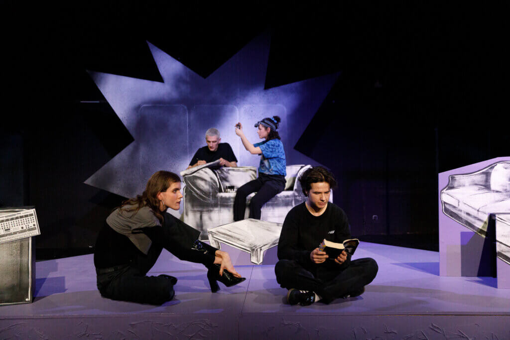 Vier Darsteller*innen auf einer Bühne, zwei im Vordergrund sitzend, zwei im Hintergrund auf einer Bank