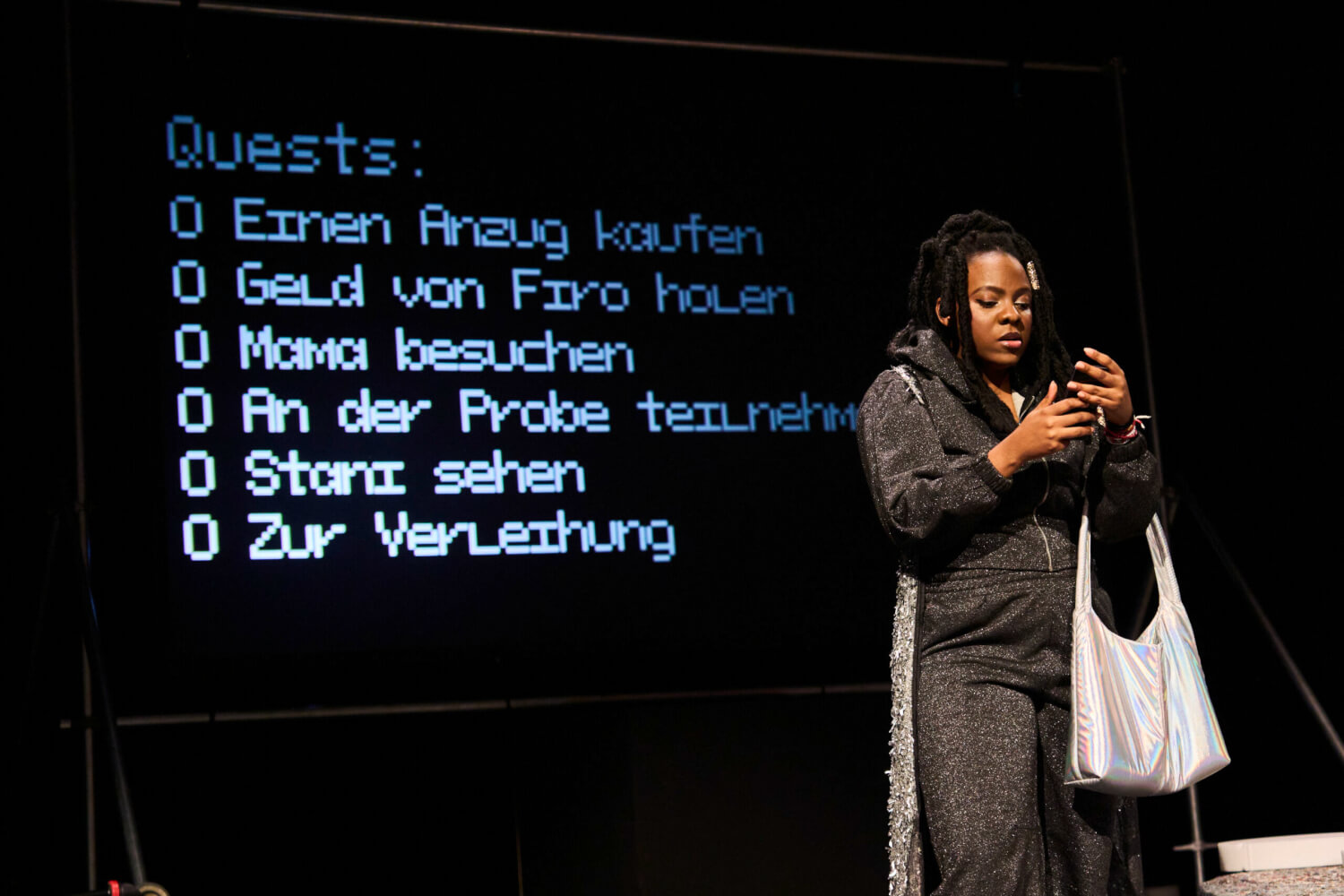 Aufführungsfoto von „Keine Aufstiegsgeschichte“ Stückentwicklung von Marco Damghani, Olivier David und Ensemble. Eine Frau im schwarz-glitzerndem Jogginganzug schaut auf ihr Handy. Über dem linken Arm trägt sie eine perlmuttfarbene Tasche. Im Hintergrund eine Leinwand mit Schrift.