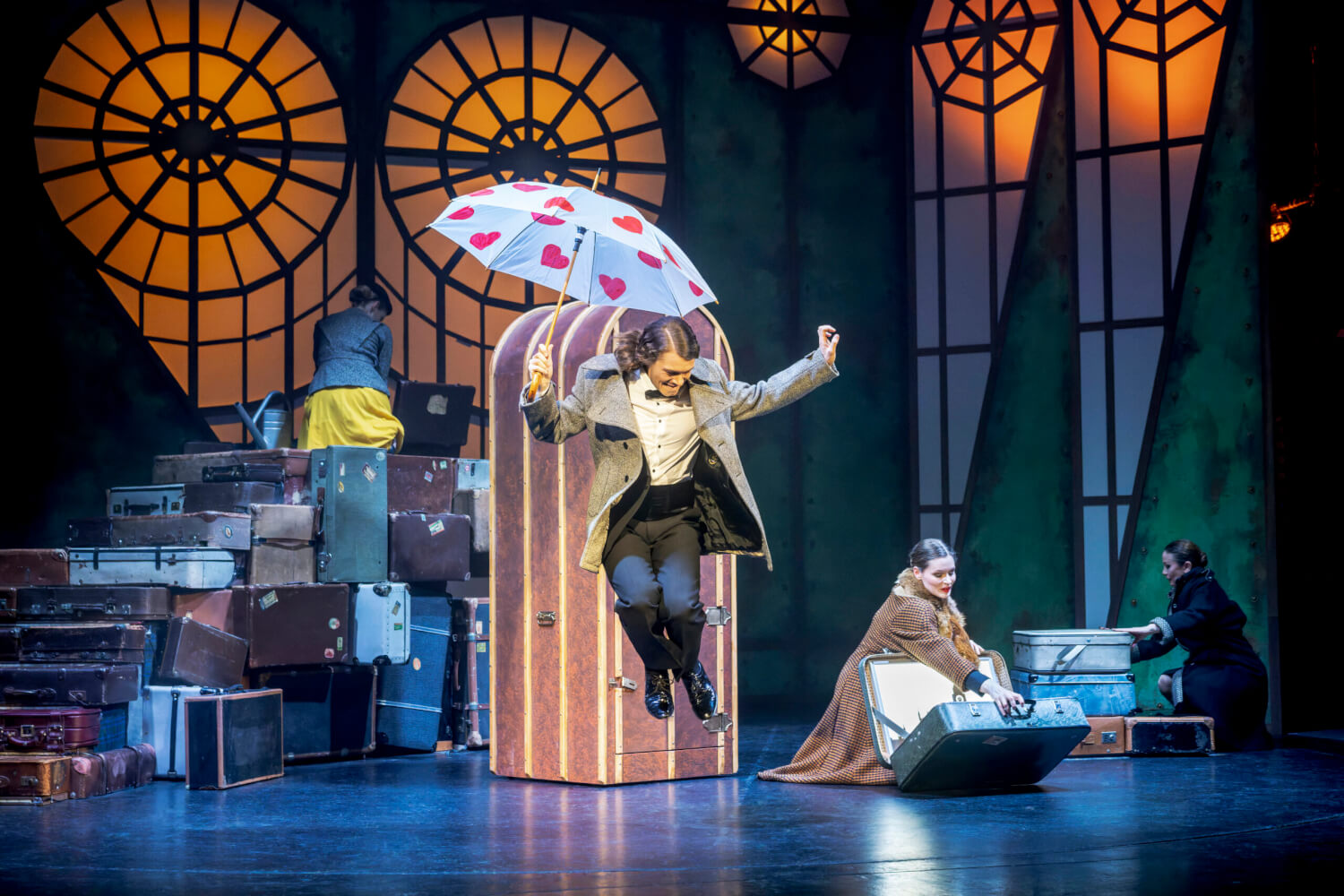 Aufführungsfoto von „Mein Freund Bunbury“ von Gerd Natschinski am Eduard-von-Winterstein Theater Annaberg-Buchholz. Ein Mann mit Herzchen-Regenschirm springt in die Luft. Im Hintergrund gestapelte Koffer und zwei Frauen, die Koffer zuklappen.