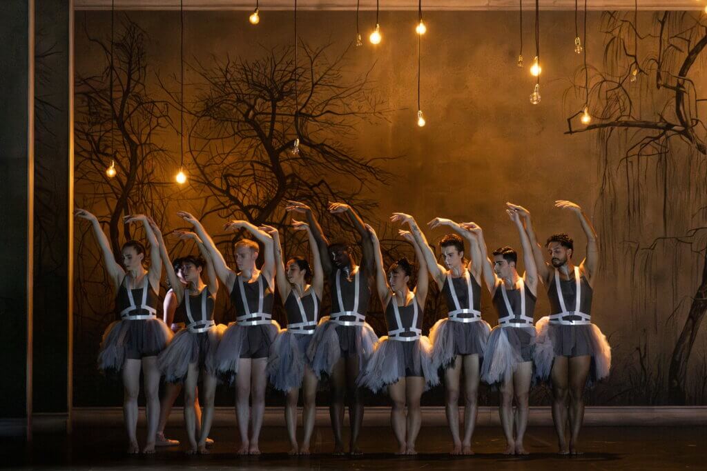 Zehn Tänzer:innen in Formation nebeneinander mit gestreckten Armen. Im Hintergrund schwarze Bäume und über der Bühne Glühbirnen.