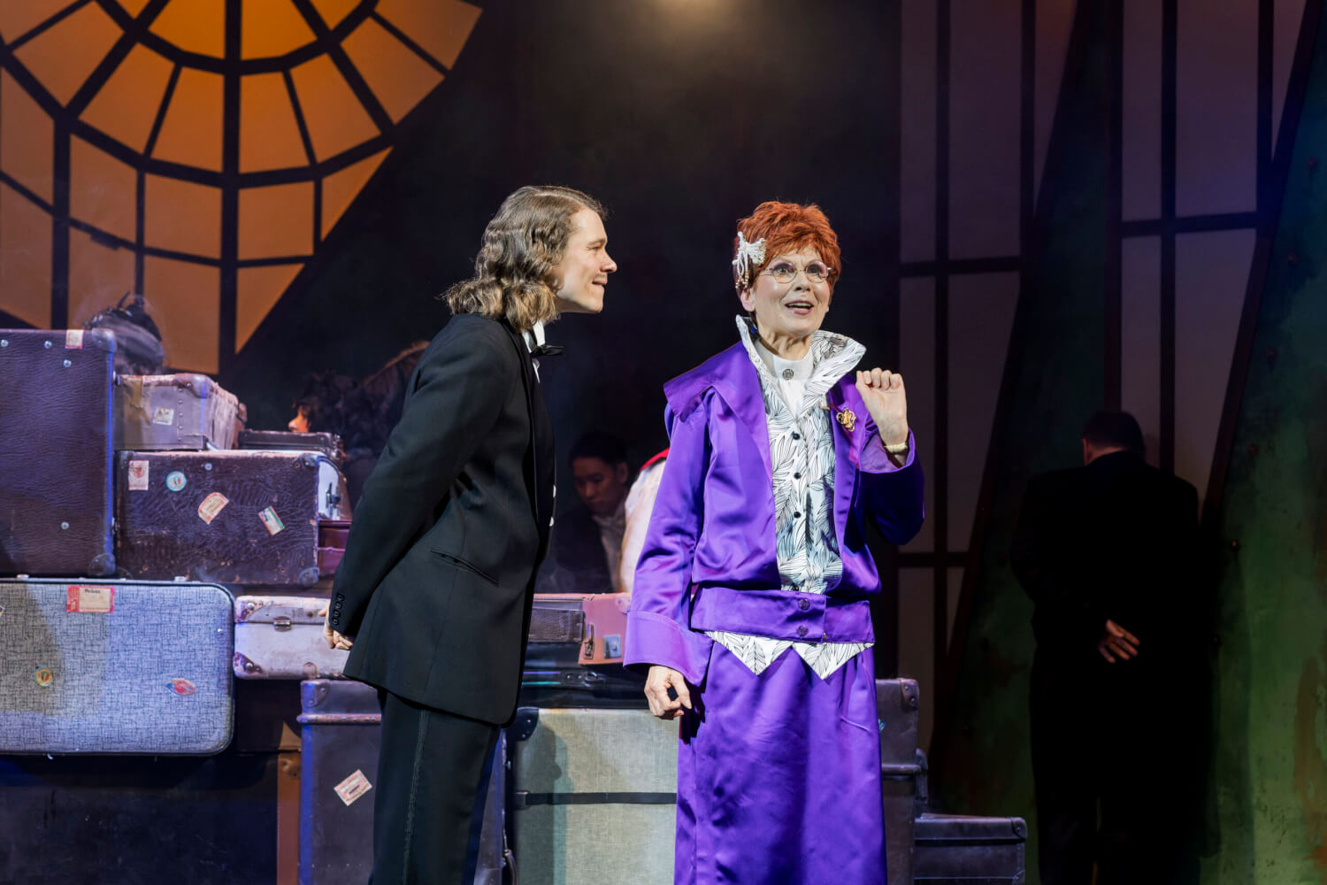 Aufführungsfoto von „Mein Freund Bunbury“ von Gerd Natschinski am Eduard-von-Winterstein Theater Annaberg-Buchholz. Im Hintergrund gestapelte Koffer. Ein Mann im schwarzen Anzug guckt eine ältere Frau seitlich sehr intensiv an, während sie in einem lila Anzug nach vorne spricht.