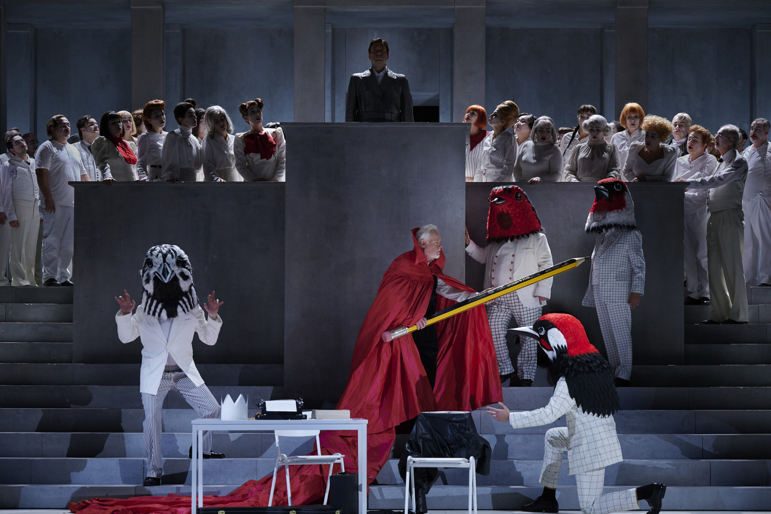 Ein Darsteller steht mit einem übergroßen Bleistift vor einem Podest und sieht aus, als würde er sich damit wehren. Um ihn herum stehen Personen mit Vogelmasken über den Köpfen. Im Hintergrund schaut der Opernchor dabei zu.