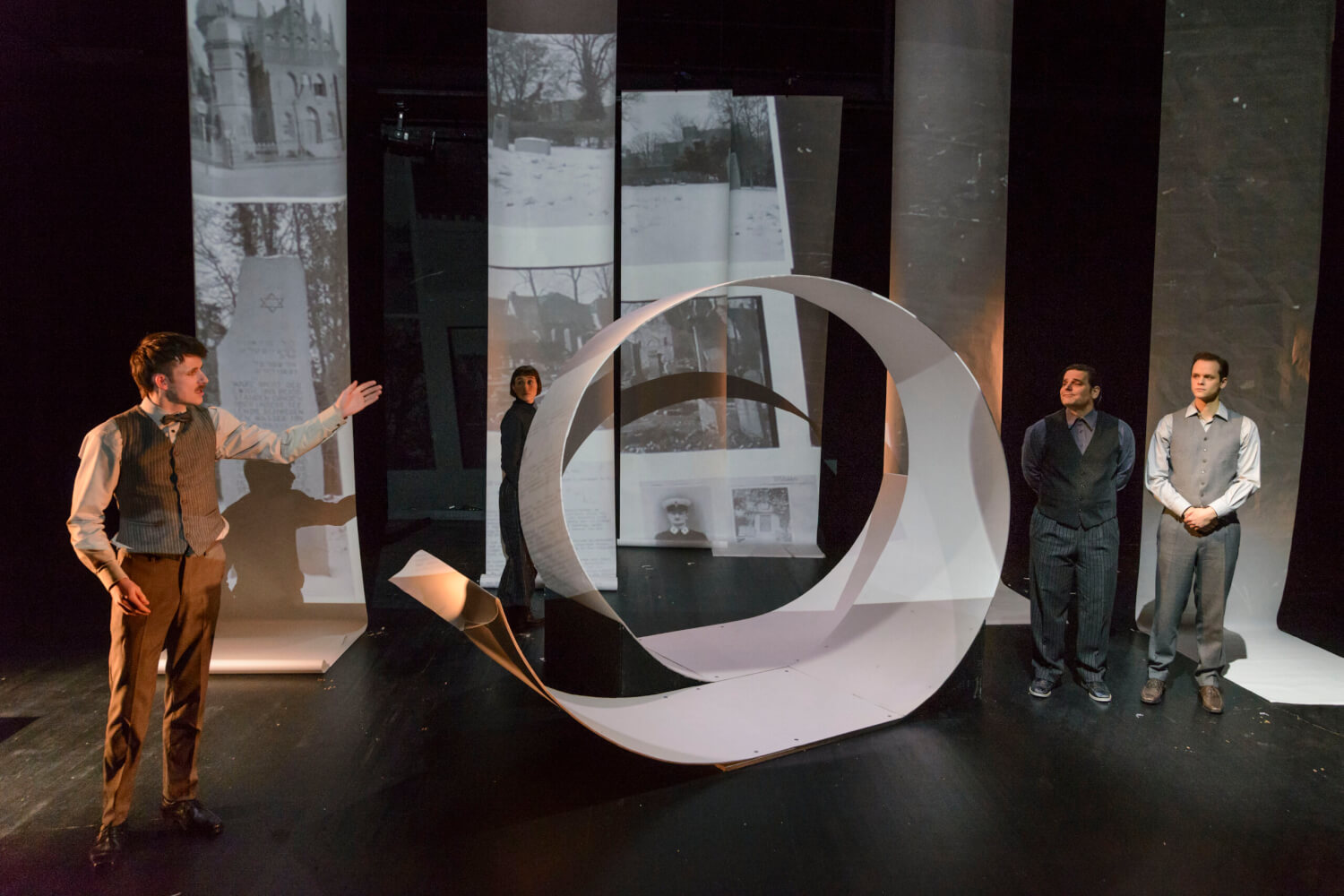 Aufführungsfoto von „Anfang – kein Ende“ von Juliane Hendes am Theater Lüneburg. In der Mitte eine metallene Spirale. Zwei Männer stehen rechts davon. Eine Frau dahinter und ein Mann links davon, der auf die zwei Männer zeigt. Im Hintergrund schwarz-weiß Fotos auf Leinwänden.