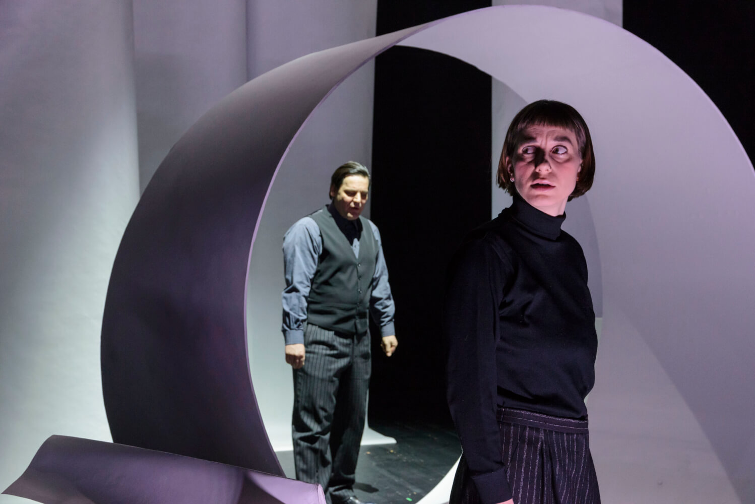 Aufführungsfoto von „Anfang – kein Ende“ von Juliane Hendes am Theater Lüneburg. Eine Frau mit kurzen Haaren im Vordergrund. Im Hintergrund steht ein Mann im Anzug in einer großen Spirale.