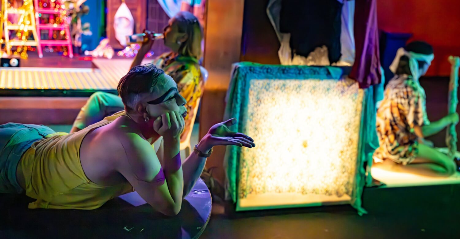 Gastspiel beim Oerol Festival auf der Insel Terschelling: „HOLOBIONT” von Boogaerdt/VanderSchoot. Eine geschminkte Person liegt auf dem Bauch, den Kopf auf die Hand gestützt, die Lippen gespitzt, die andere Hand wie zu einem Handkuss. Dahinter ein beleuchtetes Quadrat.