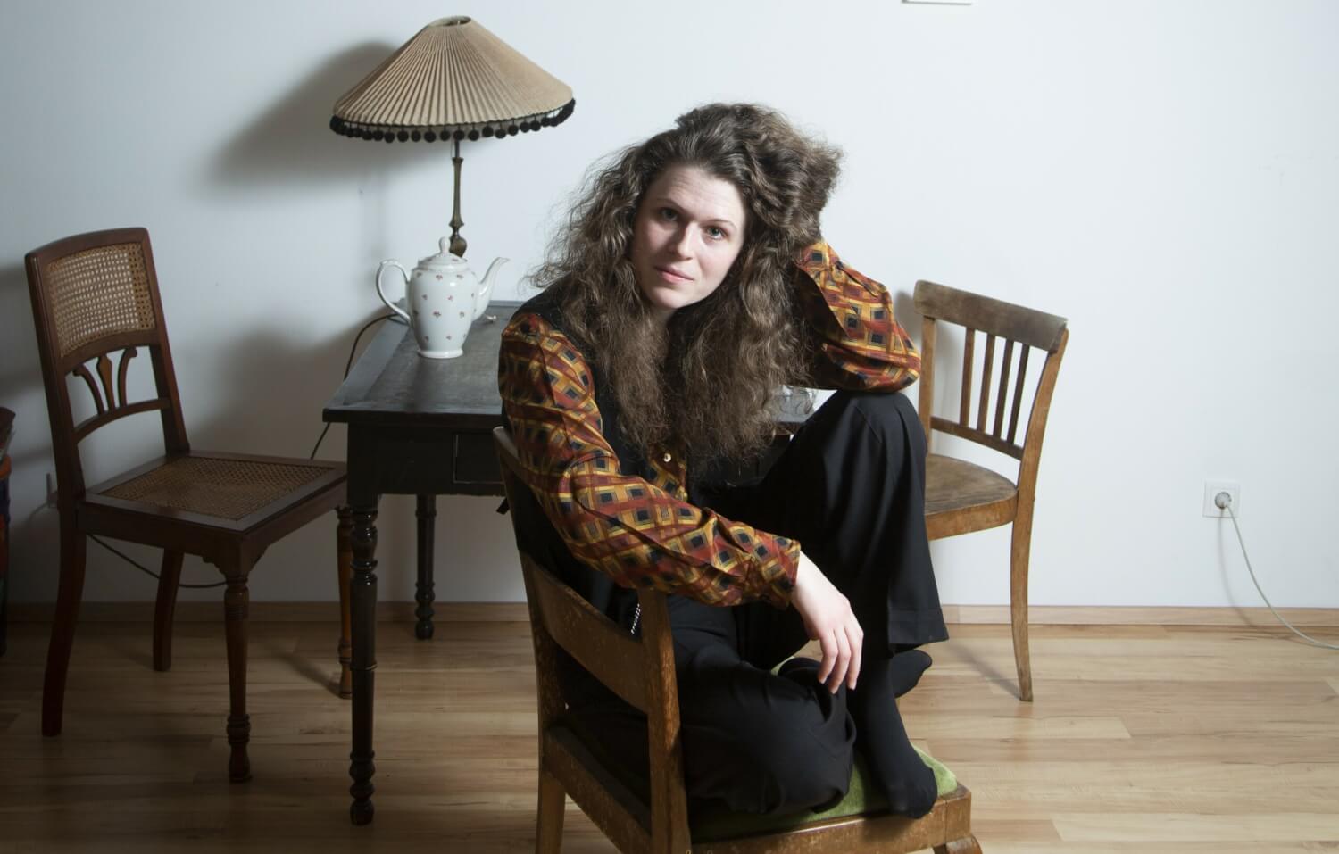 Cosmea Spelleken in ihrer Wiener Wohnung. Sie sitzt auf enem Stuhl, die Beine auf der Sitzfläche, die Hand greifen in die Haare. Im Hintergrund ein kleiner Holztisch mit zwei weiteren Stühlen. AUf dem Tisch eine Tischlampe mit Schirm und eine Porzellanteekanne.