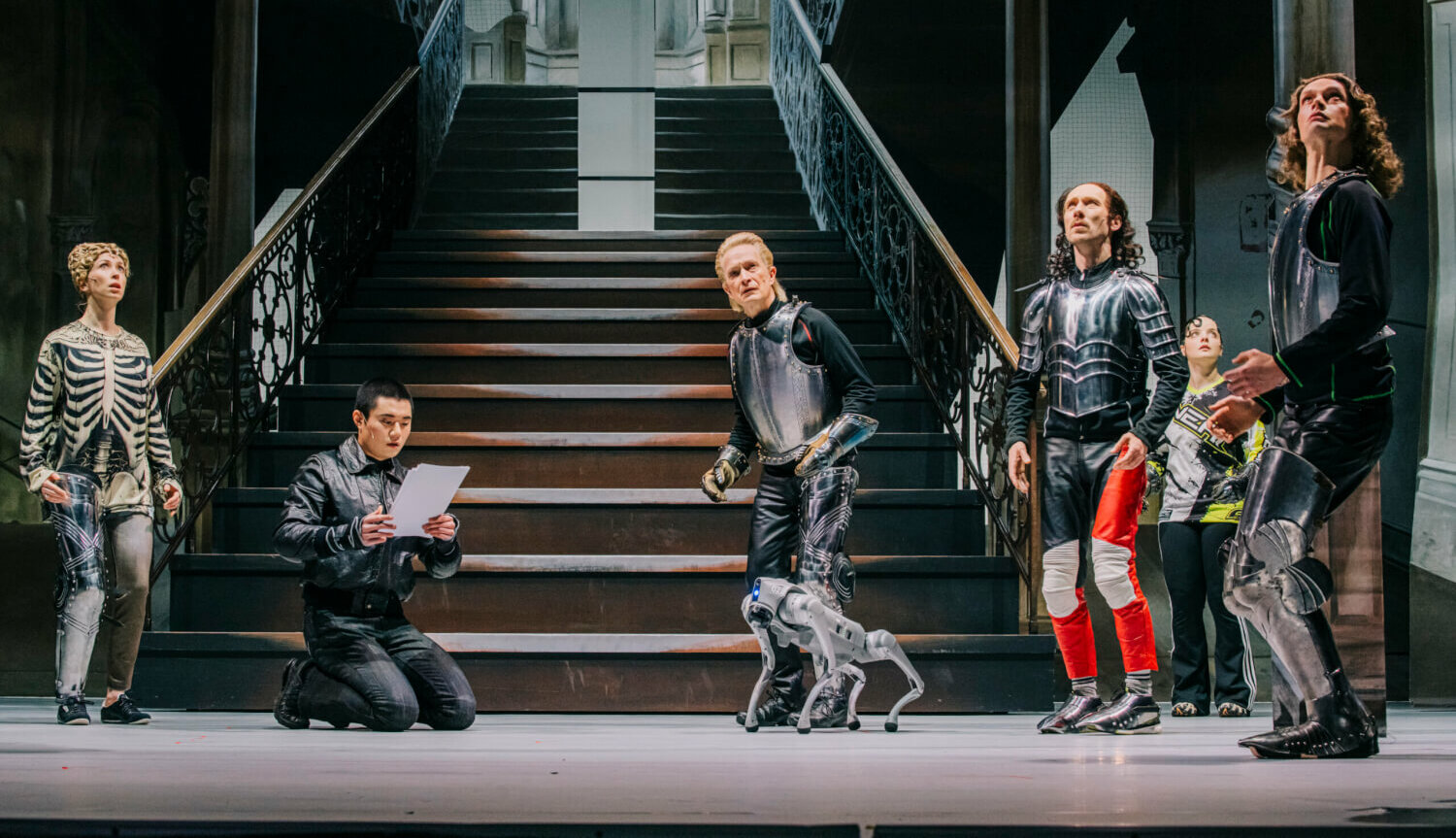 eine Bühne mit mehreren starr verharrenden Darsteller:innen und einem Robodog, im Hintergrund ein Treppenaufgang