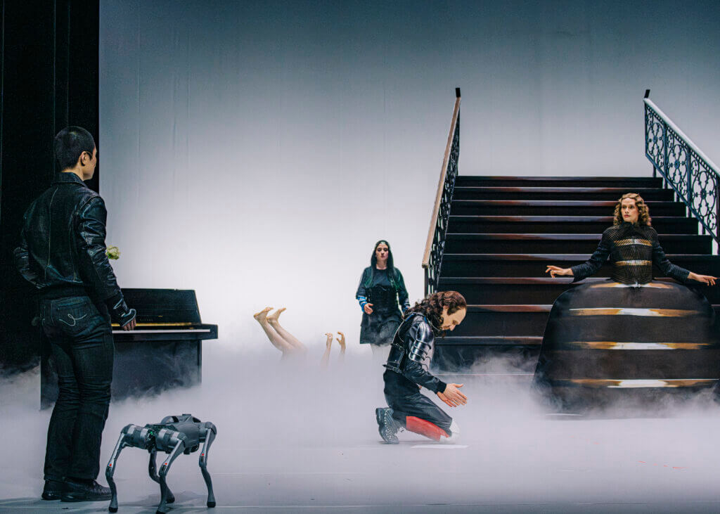 Eine am Boden benebelte Bühne mit mehreren Darstellenden, einer Treppe im Hintergrund, am vorderen Bühnenrand ein Roboterhund
