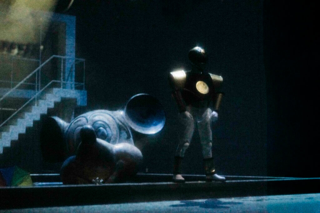 Die Bühne ist in düsteres Licht getaucht. Eine Person steht breitbeinig auf der Rechten Seite. Dahinter links liegt ein überdimensionaler Mickey Mouse-Kopf.