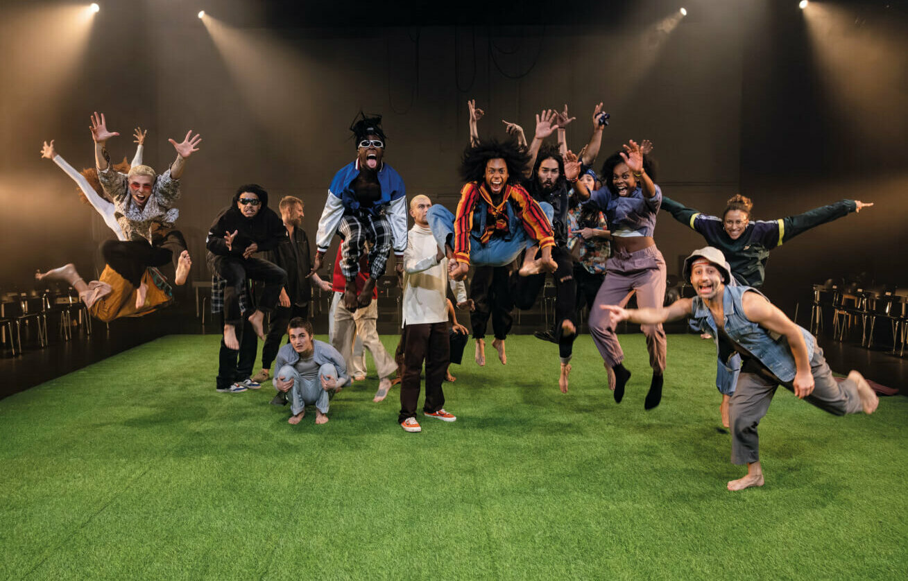 Eine diverse Tanzgruppe springt synchron und energiegeladen auf einer beleuchteten Bühne mit grünem Boden.