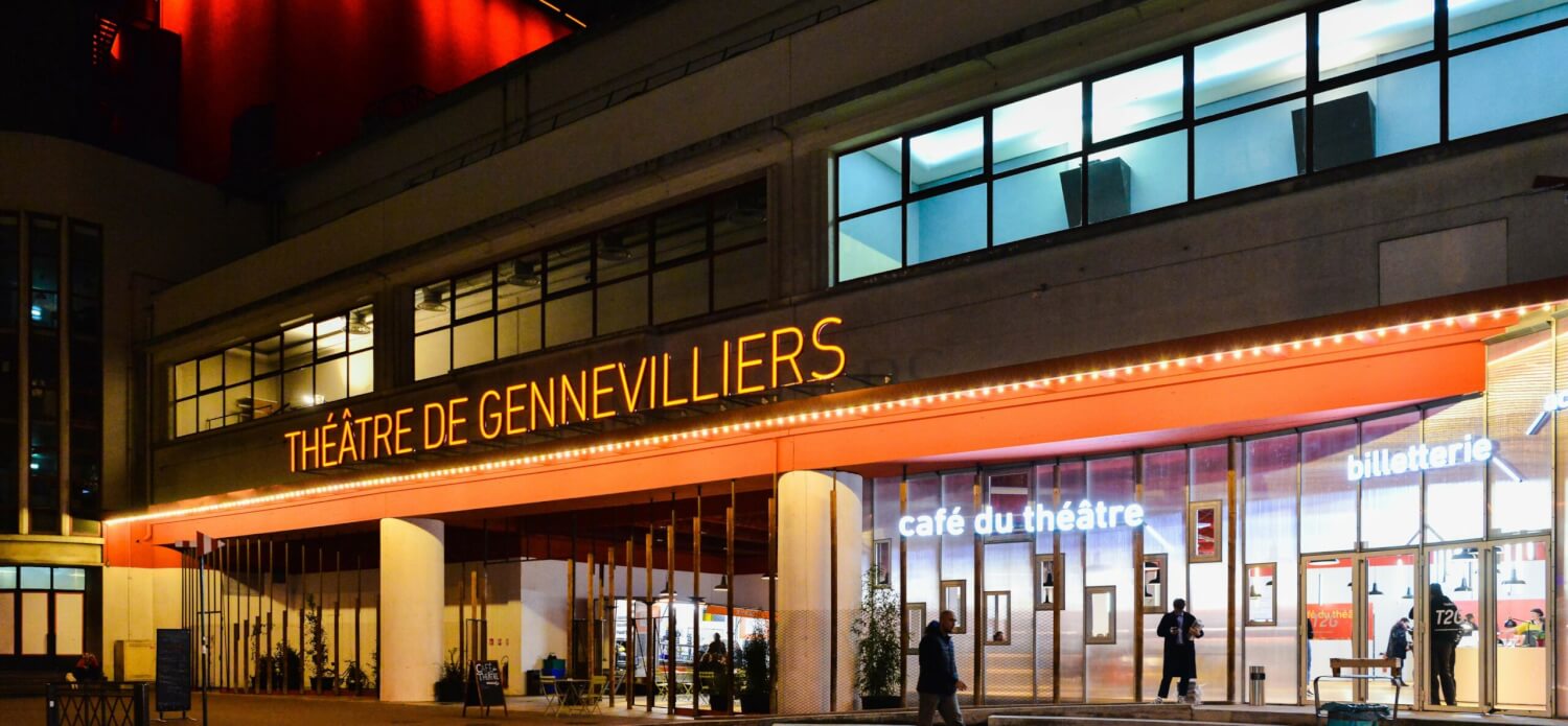 Théâtre de Gennevilliers. Die Fassade bei Nacht, der Schnürboden rot angestrahlt, die Lettern des Theaters ebenfalls beleuchtet.