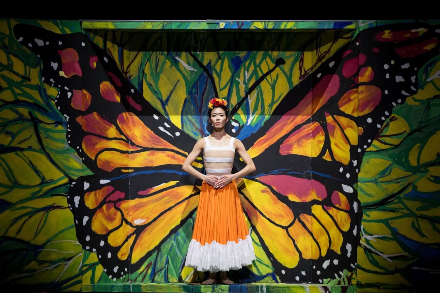 Aufführungsfoto von „Frida“ von Annabelle Lopez Ochoa am Theater Dortmund. Eine Tänzerin steht vor einer bunten Leinwand. Die Hände auf ihren Bauch gelegt. Zusammen mit der Leinwand wird die Tänzerin zu einem bunten Schmetterling.