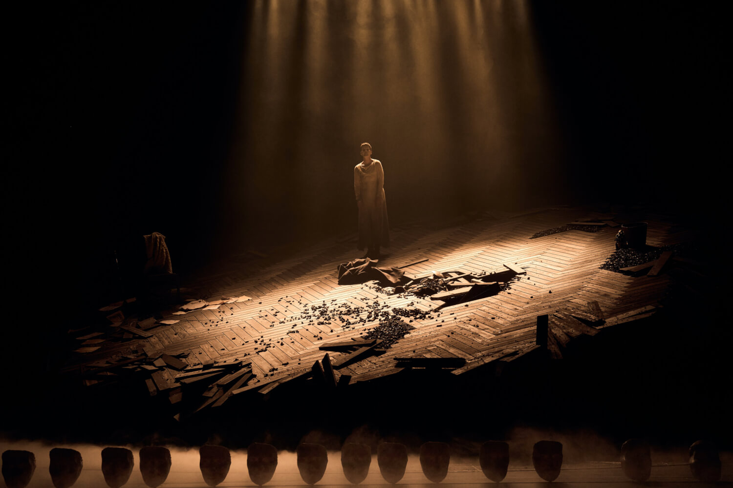 „Kassandra“ von Mathis Nitschke, Stefan Behrisch / nach Christa Wolf, mit Christina Huckle. Auf einer schrägen Parkett-Bühne steht in einem Lichtkegel eine Frau. Das Parkett ist stellenweise zerstört und mit Erde bedeckt.