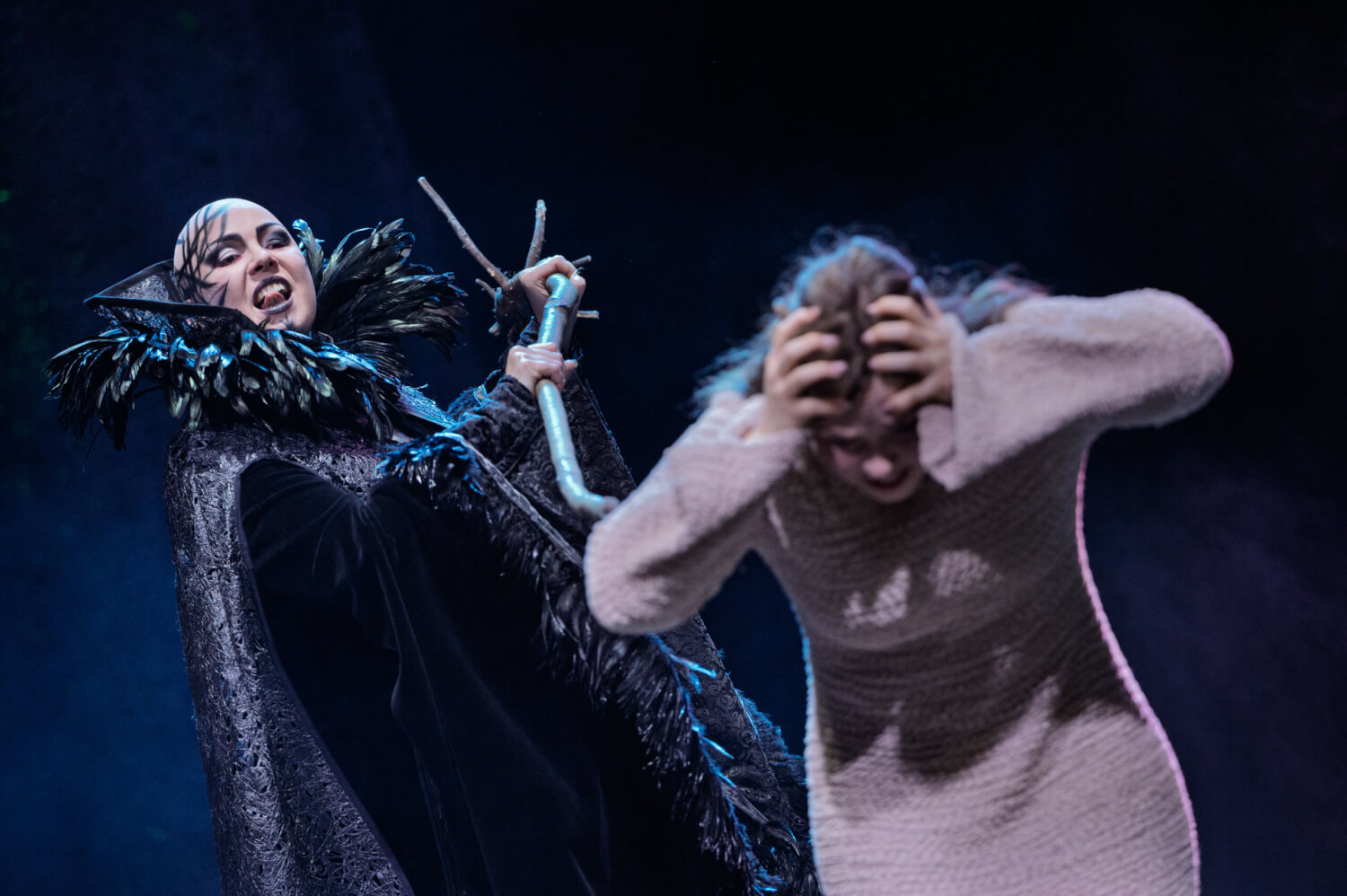 Aufführungsfoto von „Königskinder“ von Engelbert Humperdinck am Theater und Orchester Neubrandenburg Neustrelitz. Eine Frau im Vordergrund greift sich gebückt an den Kopf. Hinter ihr steht eine Hexe mit einem Stock in der Hand.