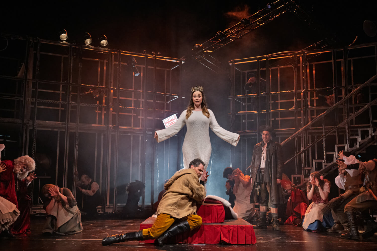 Aufführungsfoto von „Königskinder“ von Engelbert Humperdinck am Theater und Orchester Neubrandenburg Neustrelitz. Ein Metallgestell im Hintergrund. Im Vordergrund mittig eine Frau mit Krone und weißem Kleid steht auf einer Erhöhung. Ihr zu Füßen ein Mann, der sich an sie lehnt.