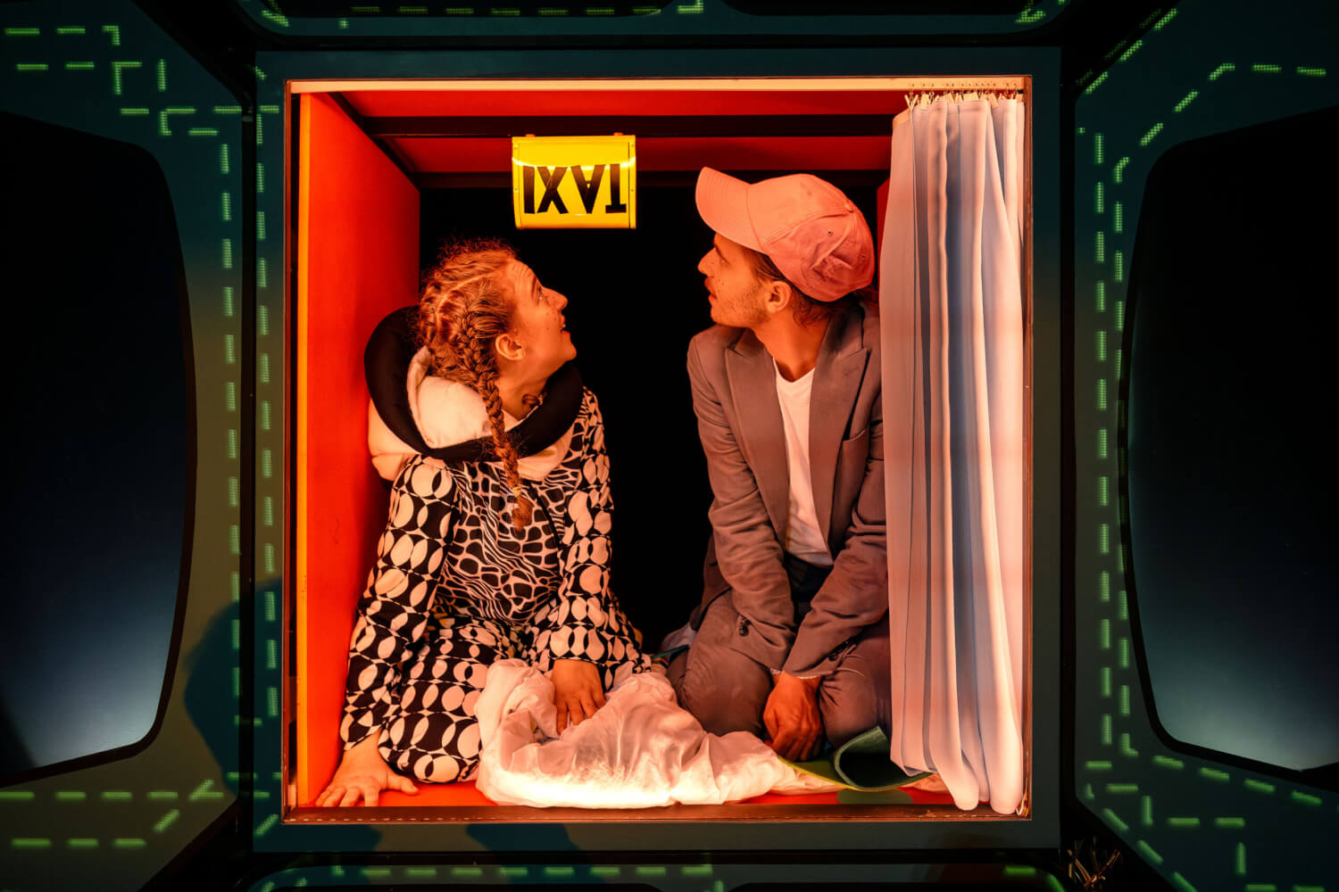 Anna Lehotská und Jakob Ferdinand Lenk sitzen in einer kleinen, beleuchteten Kiste an der ein Schild mit Aufschrift "Taxi" verkehrt herum hängt.