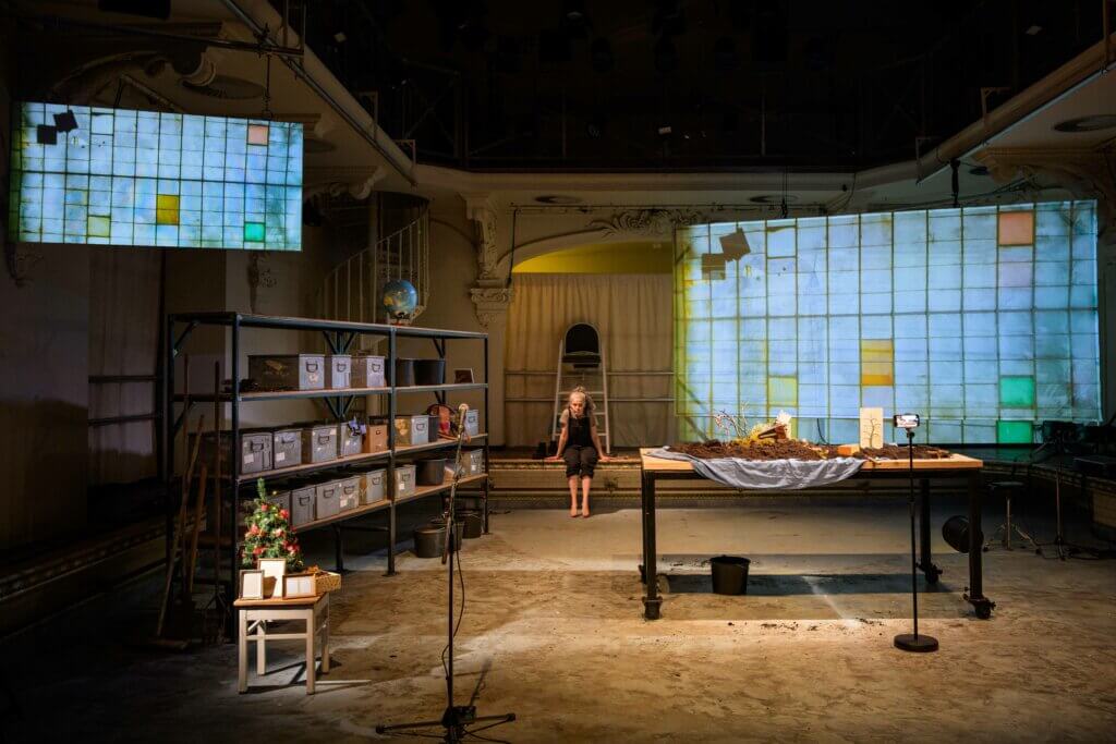 Die Bühne ist als Halle mit großen Fenstern, gefüllten Regalen und einem frei stehenden Arbeitstisch mit Gartenutensilien ausgestattet.