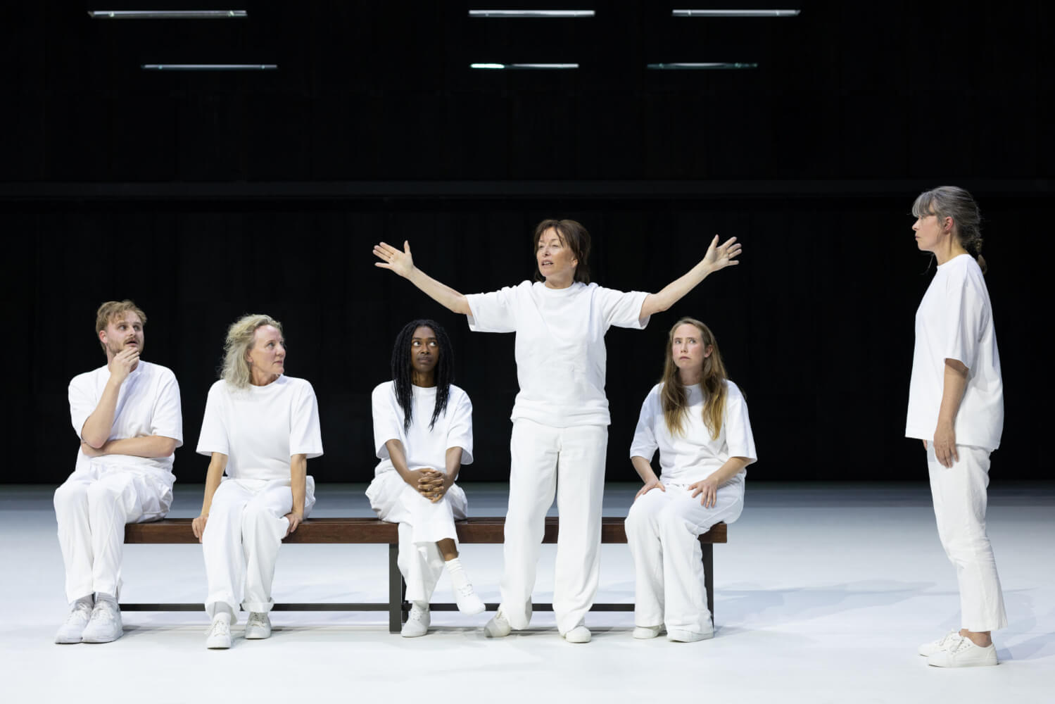„Tijd voor geluk“ am Internationaal Theater Amsterdam. Vier Personen, mit weißen Hosen und weißen T-shirts, sitzen auf einer Bank. Eine Frau steht vor der Bank und hebt die Arme empor. Eine andere Frau steht rechts daneben mit hängenden Armen.