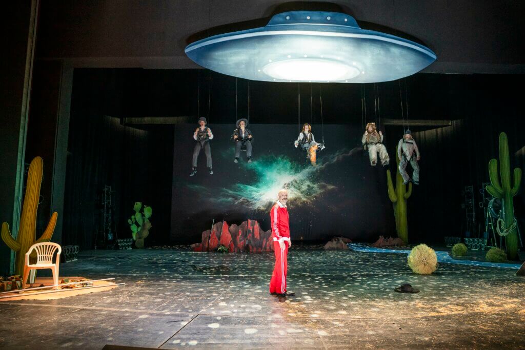 Moritz Grove steht vorne in der Mitte der Bühne im Trainingsanzug. Hinter ihm baumelt der Rest des Ensembles unter einem UFO.