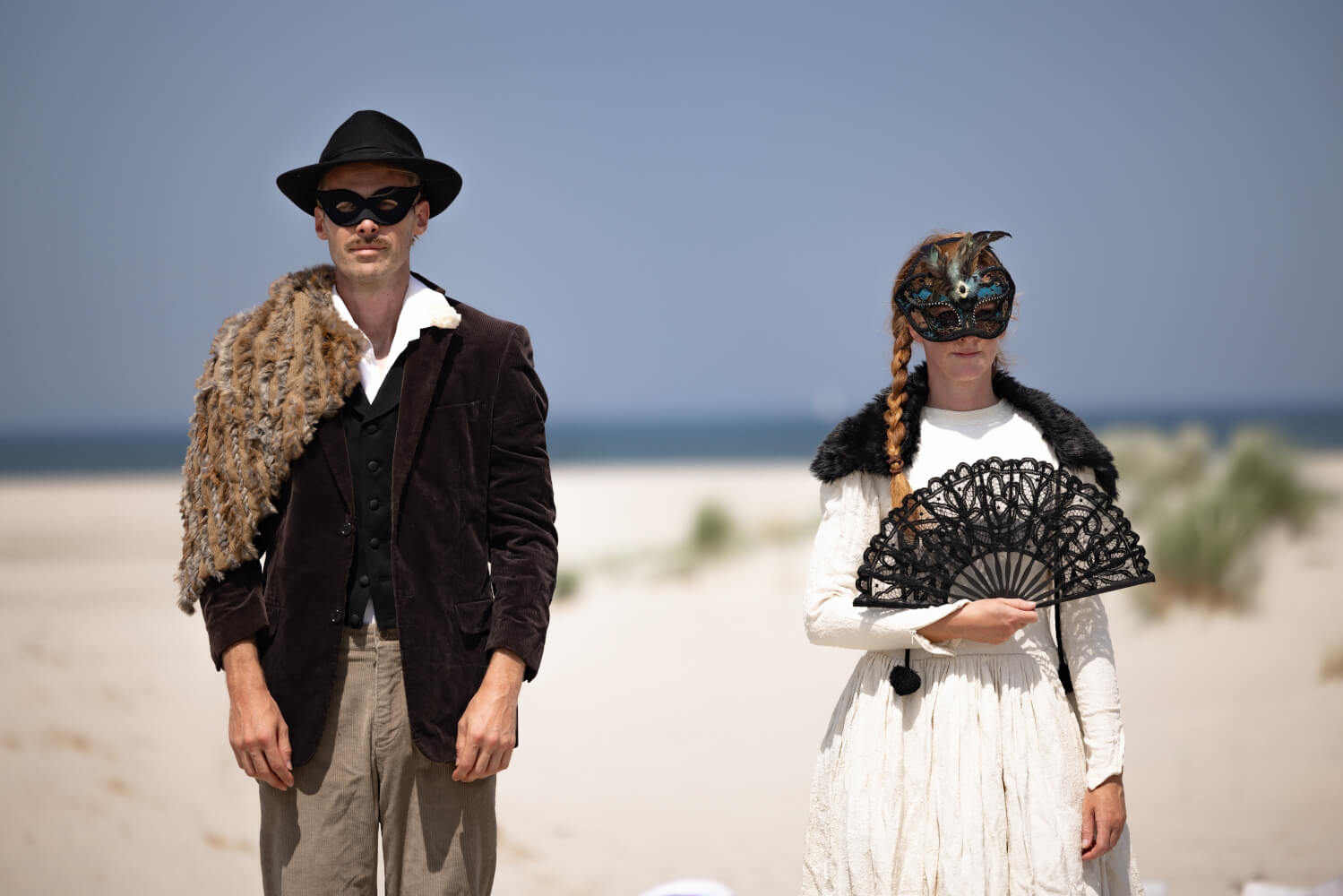 Gastspiel beim Oerol Festival auf der Insel Terschelling: „Catwalk” von Zwermers. Ein Mann und eine Frau stehen nebeneinander, tragen schwarze Masken. Im Hintergrund Strand und das Meer. Sie hält einen schwarzen Fächer in der Hand.
